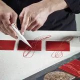 Mains créatives réalisant une décoration en chocolat facile et rapide sur des carrés rouges brillants.
