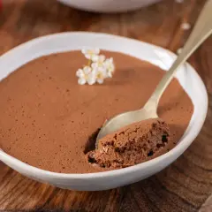 Une cuillère d'une mousse au chocolat compacte et onctueuse, garnie de petites fleurs blanches, dans un bol blanc sur fond de bois.