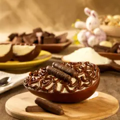 Un œuf au chocolat garni de crème et de bâtonnets de chocolat, prêt à être dégusté. Des morceaux de chocolat et un lapin de Pâques en arrière-plan.