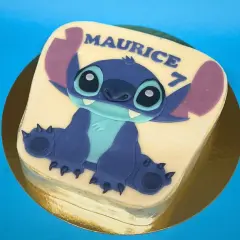 Gâteau thème Stitch pour Maurice, qui fête ses 7 ans. Le personnage bleu est assis sur un glaçage blanc.