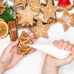 Mains décorant un biscuit de noël en forme de sapin avec du glaçage blanc. D'autres biscuits étoilés et décorations festives sont visibles.