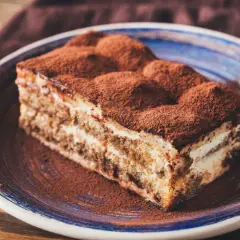 Une délicieuse part de tiramisu, généreusement saupoudrée de cacao, sur une assiette bleue.