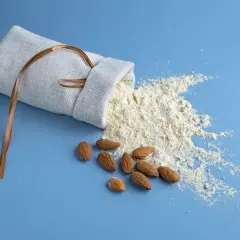 Un sac en lin renversé laisse échapper de la farine d'amande ou poudre d'amande, avec quelques amandes entières à proximité.