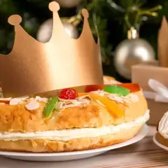 Un roi porte une couronne dorée sur un délicieux gâteau praliné, décoré de fruits confits et d'amandes. Un cadeau emballé et des décorations de Noël en arrière-plan.