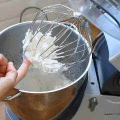 Un doigt teste la texture de la chantilly tranchée sur le fouet d'un robot pâtissier.