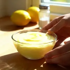 Un bol de crème au citron sans oeuf, onctueuse et jaune vif, est présenté sur un plan de travail en bois. Des citrons et une salière sont en arrière-plan.
