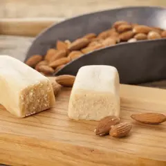 Deux blocs de pâte d'amande maison sur une planche en bois, avec des amandes dans une pelle en arrière-plan.