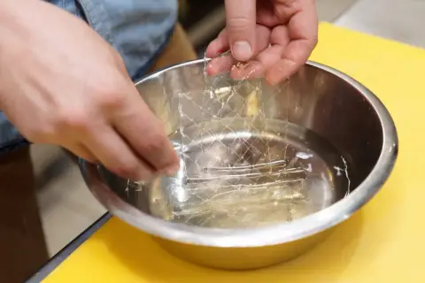 Mains trempant une feuille de gélatine dans l'eau froide. C'est la première étape pour savoir comment utiliser la gélatine en feuille.