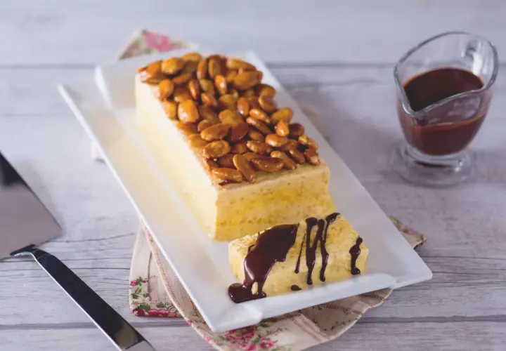 Gâteau rectangulaire glacé, garni d'amandes fruit caramélisées. Une part coupée est arrosée de sauce chocolat.