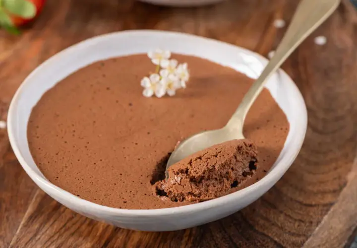 Une cuillère d'une mousse au chocolat compacte et onctueuse, garnie de petites fleurs blanches, dans un bol blanc sur fond de bois.
