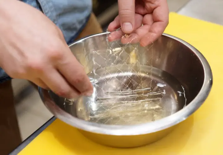 Mains trempant une feuille de gélatine dans l'eau froide. C'est la première étape pour savoir comment utiliser la gélatine en feuille.
