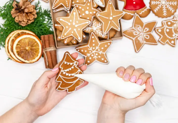 Mains décorant un biscuit de noël en forme de sapin avec du glaçage blanc. D'autres biscuits étoilés et décorations festives sont visibles.