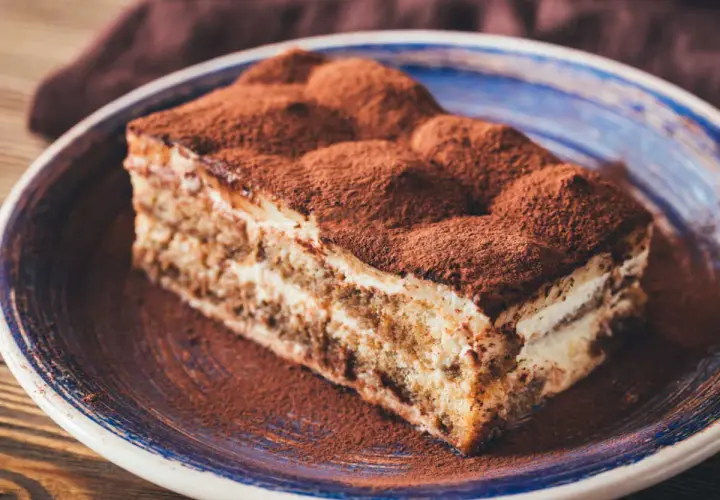 Une délicieuse part de tiramisu, généreusement saupoudrée de cacao, sur une assiette bleue.