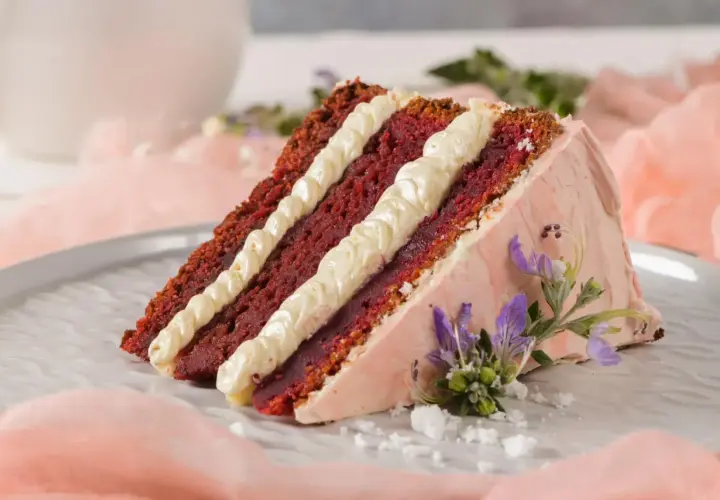 Tranche de gâteau Red Velvet avec une garniture molly cake crémeuse, décorée de fleurs violettes et de sucre glace.