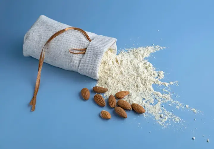 Un sac en lin renversé laisse échapper de la farine d'amande ou poudre d'amande, avec quelques amandes entières à proximité.
