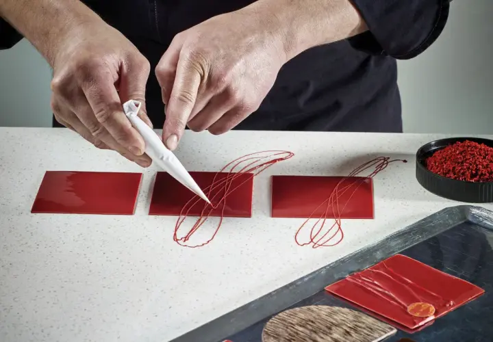 Mains créatives réalisant une décoration en chocolat facile et rapide sur des carrés rouges brillants.