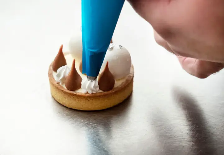Un chef utilise une poche à douille bleue pour le pochage d'une crème sur un petit gâteau.