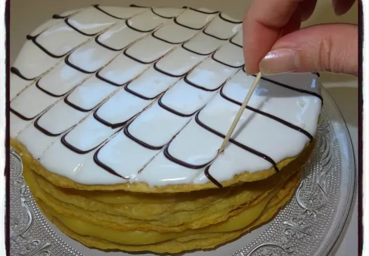 Un millefeuille rond, garni de crème pâtissière et décoré de glaçage blanc et chocolat, est finalisé avec une pique en bois.