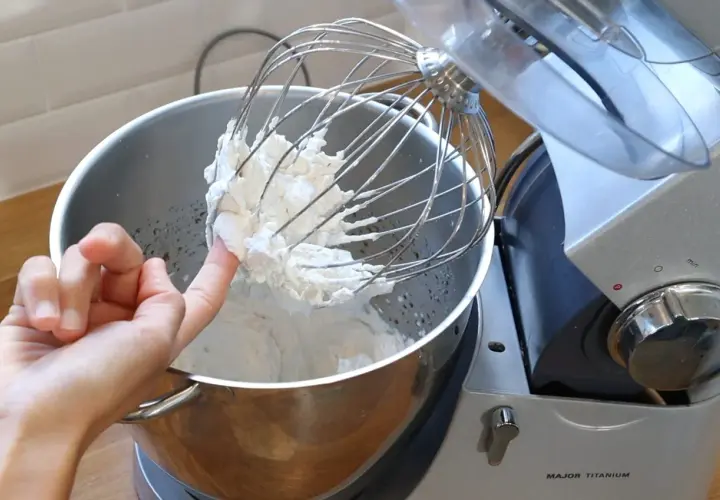 Un doigt teste la texture de la chantilly tranchée sur le fouet d'un robot pâtissier.