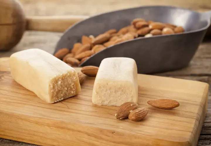 Deux blocs de pâte d'amande maison sur une planche en bois, avec des amandes dans une pelle en arrière-plan.