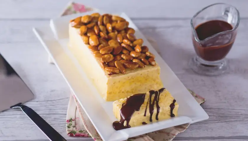 Gâteau rectangulaire glacé, garni d'amandes fruit caramélisées. Une part coupée est arrosée de sauce chocolat.