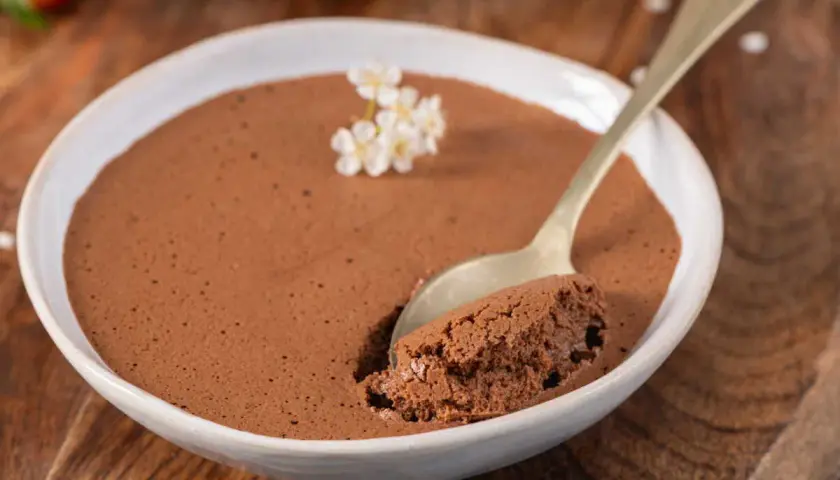 Une cuillère d'une mousse au chocolat compacte et onctueuse, garnie de petites fleurs blanches, dans un bol blanc sur fond de bois.