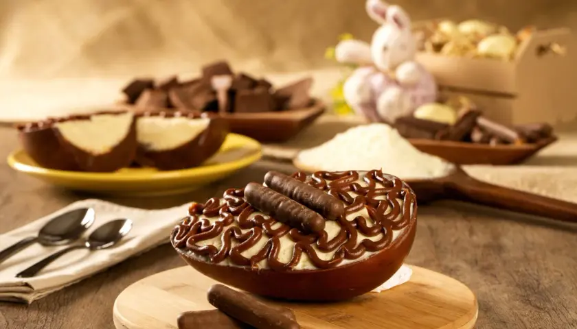 Un œuf au chocolat garni de crème et de bâtonnets de chocolat, prêt à être dégusté. Des morceaux de chocolat et un lapin de Pâques en arrière-plan.