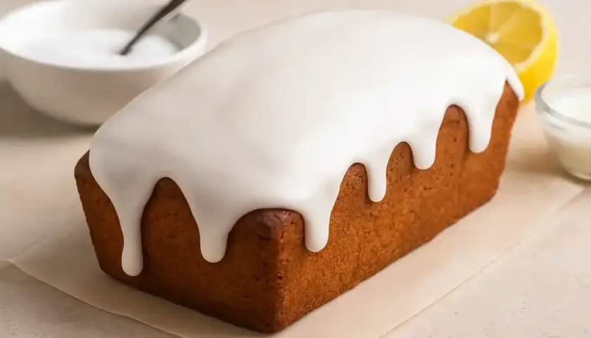 Glaçage au sucre glace et eau dégoulinant sur un cake doré. Une tranche de citron et un bol de glaçage sont à proximité.