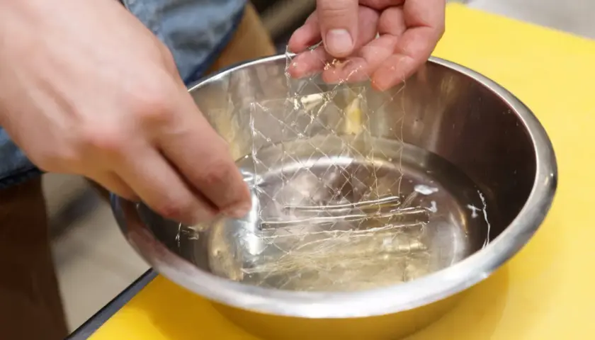 Mains trempant une feuille de gélatine dans l'eau froide. C'est la première étape pour savoir comment utiliser la gélatine en feuille.