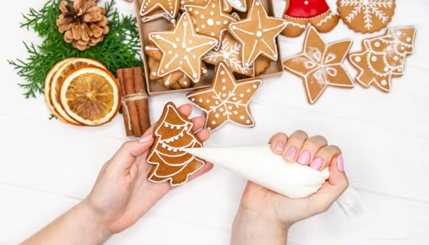 Mains décorant un biscuit de noël en forme de sapin avec du glaçage blanc. D'autres biscuits étoilés et décorations festives sont visibles.