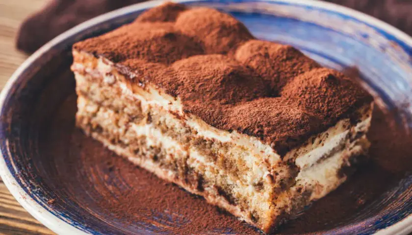 Une part généreuse de tiramisu cake, saupoudrée de cacao, repose sur une assiette bleue. Un délice italien.