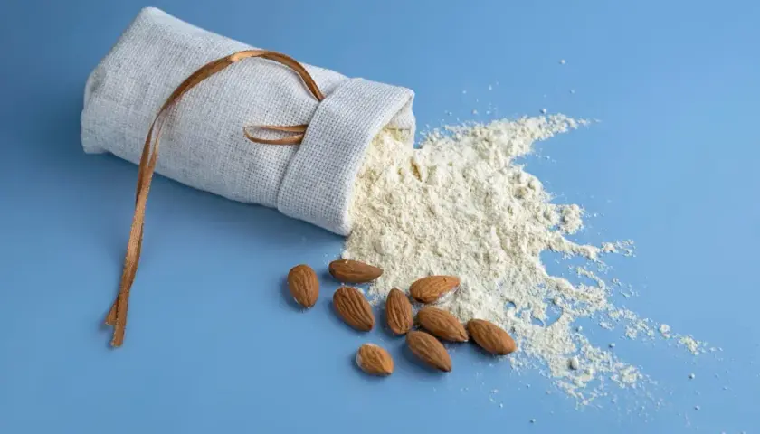 Un sac en lin renversé laisse échapper de la farine d'amande ou poudre d'amande, avec quelques amandes entières à proximité.