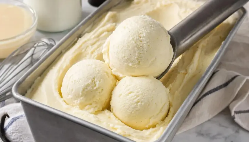 Une cuillère prélève des boules de glace à la vanille dans un bac. Une recette glace maison parfaite pour l'été.