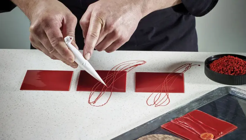 Mains créatives réalisant une décoration en chocolat facile et rapide sur des carrés rouges brillants.