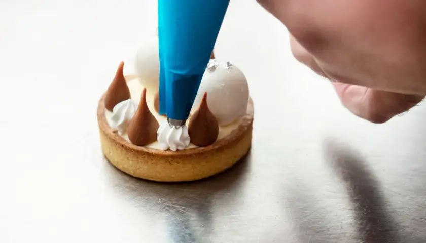 Un chef utilise une poche à douille bleue pour le pochage d'une crème sur un petit gâteau.