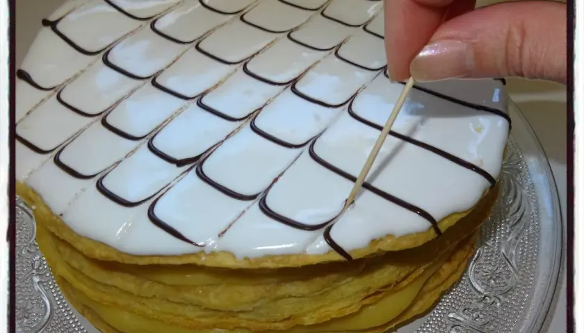 Un millefeuille rond, garni de crème pâtissière et décoré de glaçage blanc et chocolat, est finalisé avec une pique en bois.