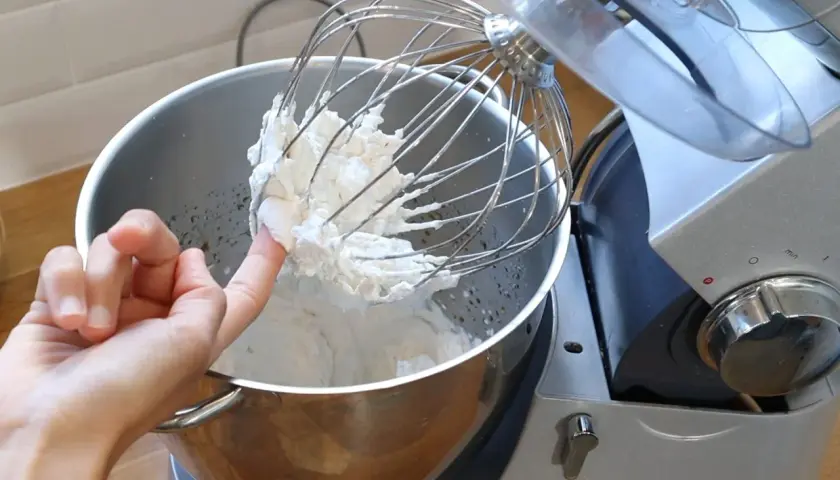 Un doigt teste la texture de la chantilly tranchée sur le fouet d'un robot pâtissier.