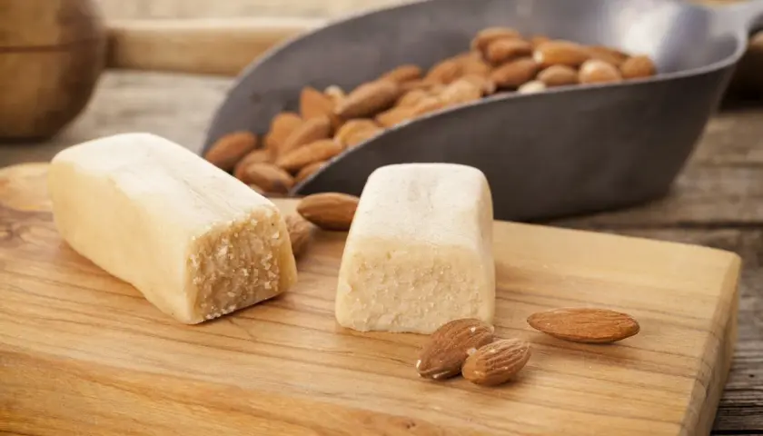 Deux blocs de pâte d'amande maison sur une planche en bois, avec des amandes dans une pelle en arrière-plan.