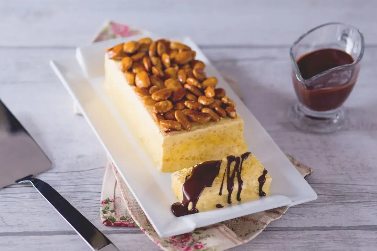Gâteau rectangulaire glacé, garni d'amandes fruit caramélisées. Une part coupée est arrosée de sauce chocolat.