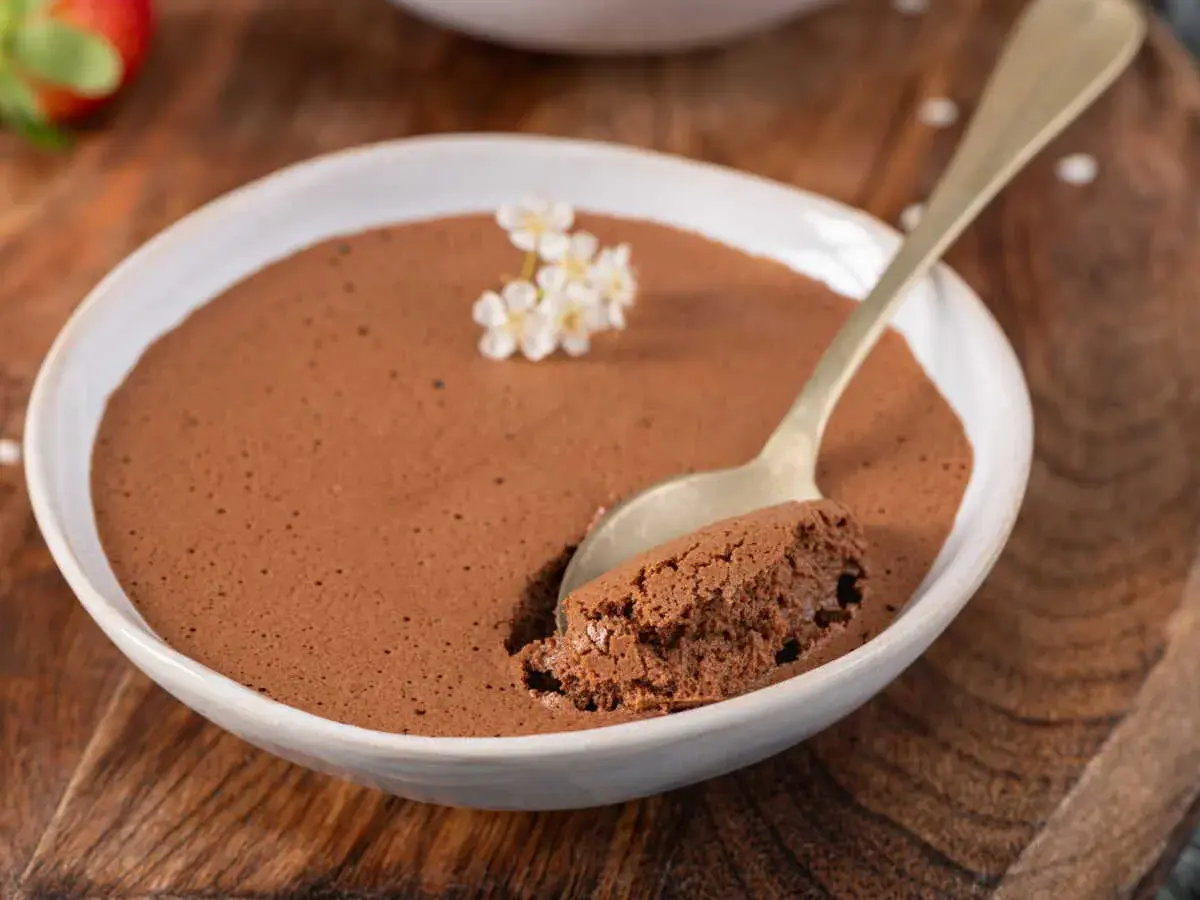 Une cuillère d'une mousse au chocolat compacte et onctueuse, garnie de petites fleurs blanches, dans un bol blanc sur fond de bois.