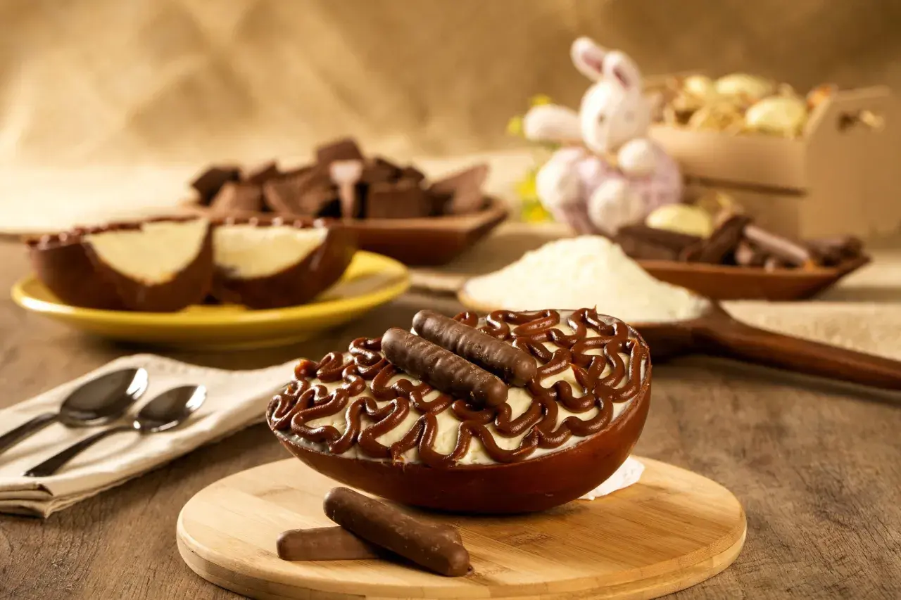 Un œuf au chocolat garni de crème et de bâtonnets de chocolat, prêt à être dégusté. Des morceaux de chocolat et un lapin de Pâques en arrière-plan.