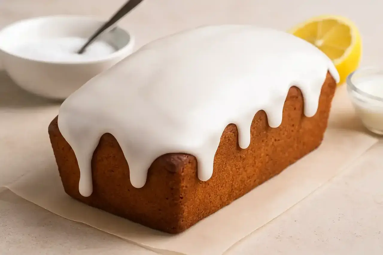 Glaçage au sucre glace et eau dégoulinant sur un cake doré. Une tranche de citron et un bol de glaçage sont à proximité.