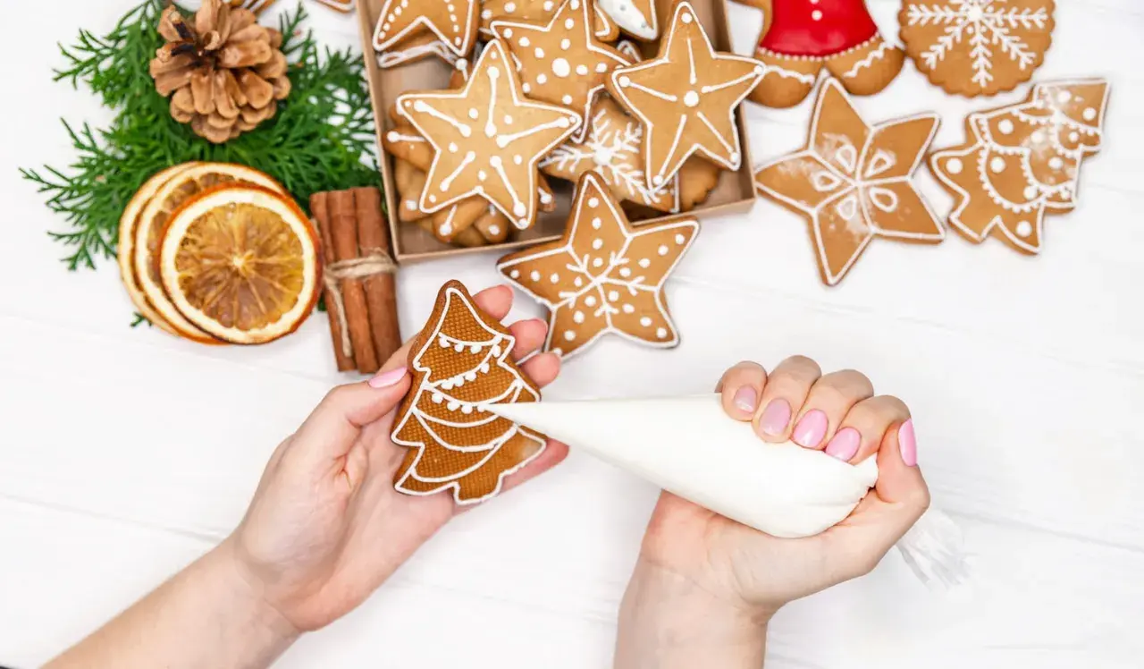 Mains décorant un biscuit de noël en forme de sapin avec du glaçage blanc. D'autres biscuits étoilés et décorations festives sont visibles.