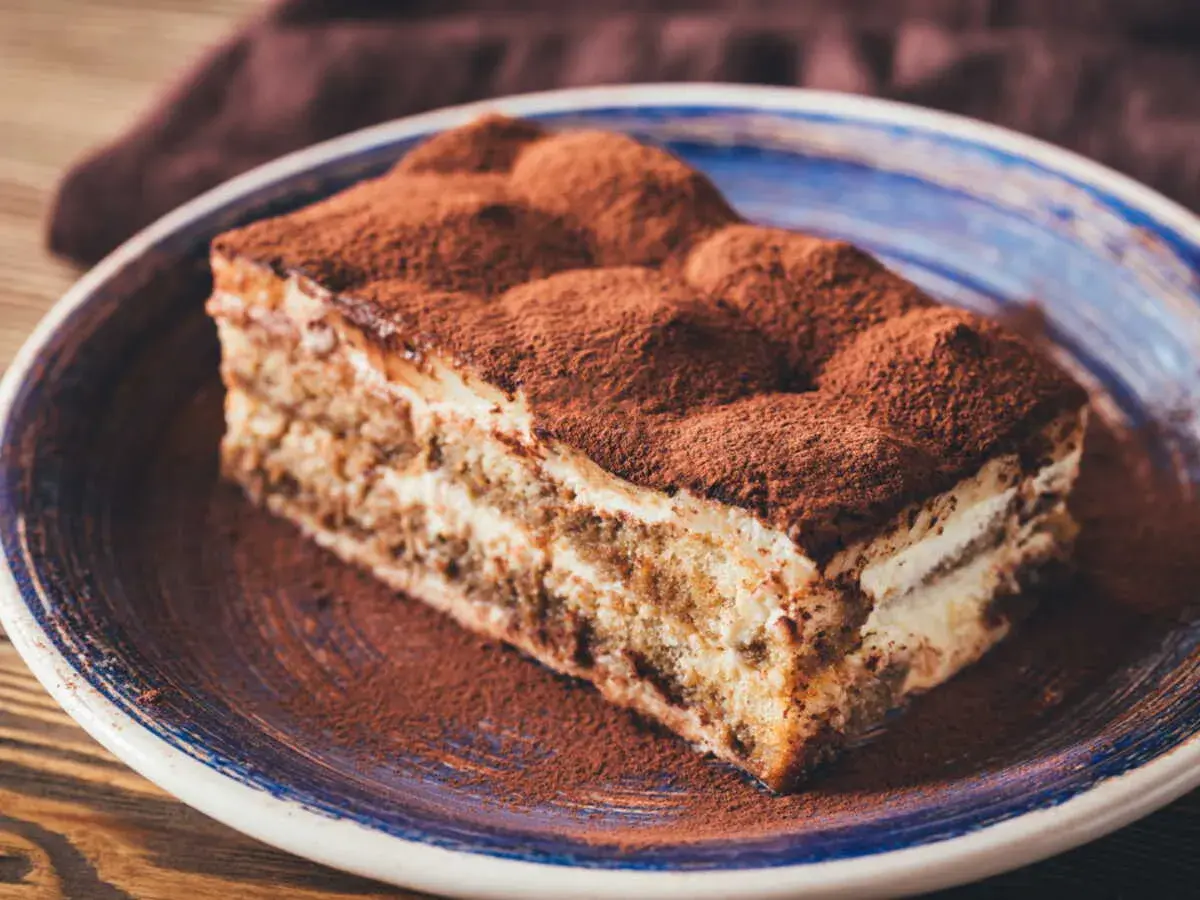Une délicieuse part de tiramisu, généreusement saupoudrée de cacao, sur une assiette bleue.