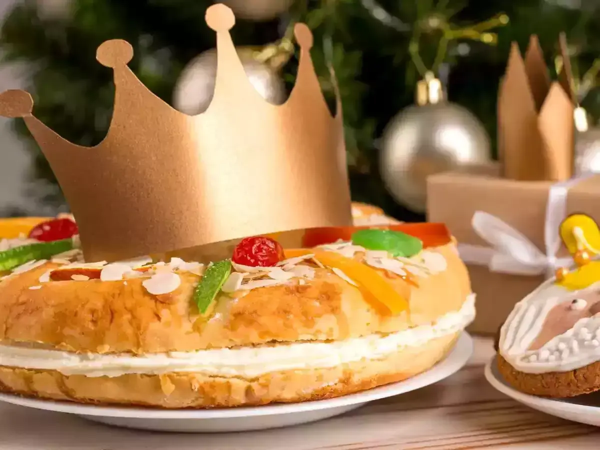 Un roi porte une couronne dorée sur un délicieux gâteau praliné, décoré de fruits confits et d'amandes. Un cadeau emballé et des décorations de Noël en arrière-plan.