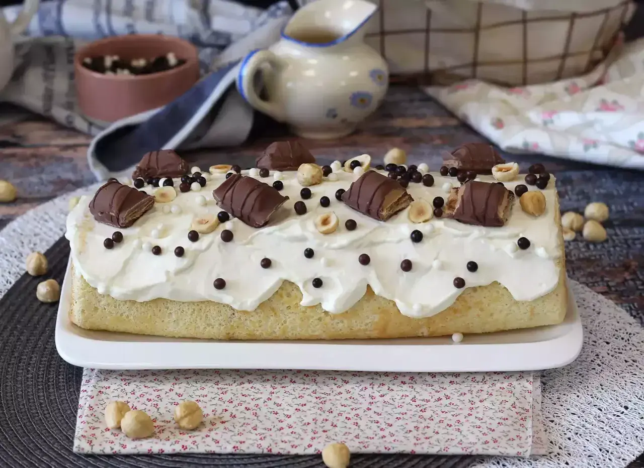 Gâteau moelleux recouvert de crème fouettée, décoré de morceaux de Kinder Bueno, noisettes et billes chocolatées. Un délice gourmand !
