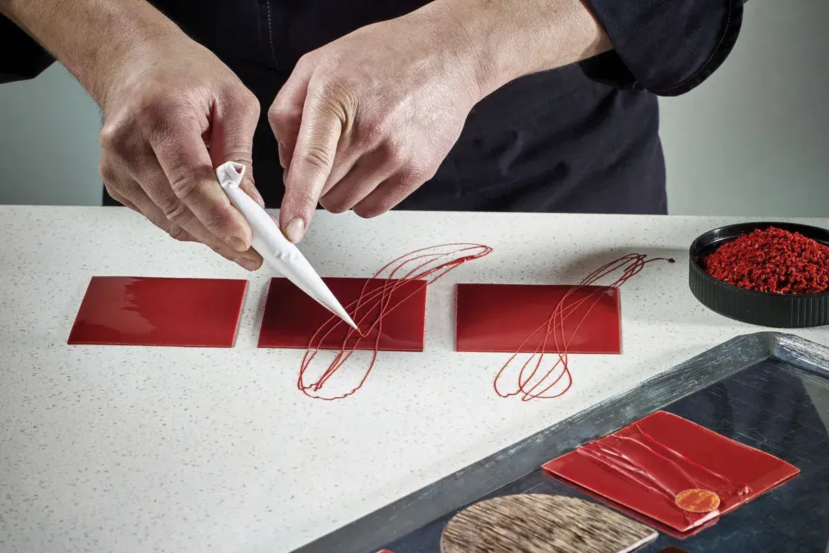 Mains créatives réalisant une décoration en chocolat facile et rapide sur des carrés rouges brillants.