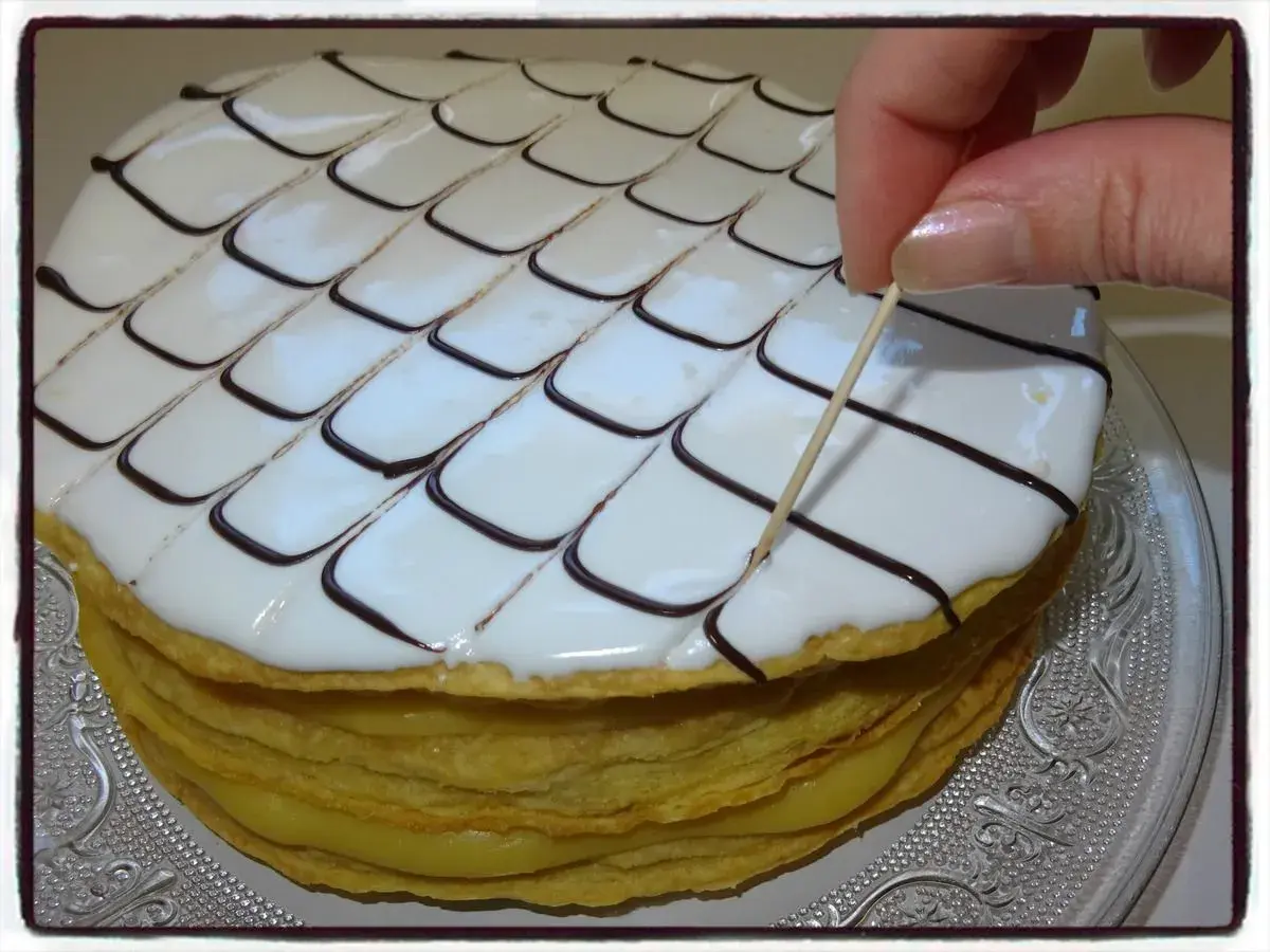 Un millefeuille rond, garni de crème pâtissière et décoré de glaçage blanc et chocolat, est finalisé avec une pique en bois.