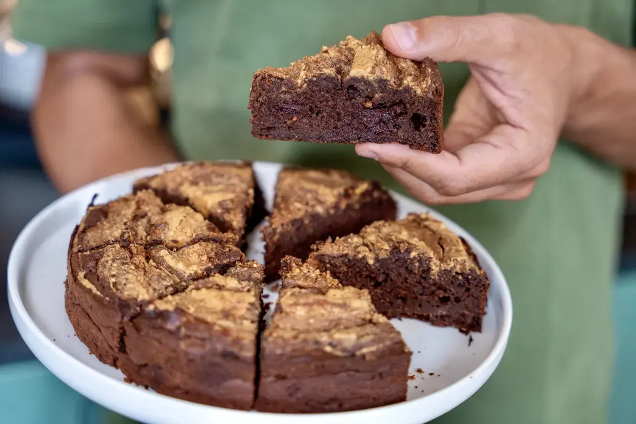 Une main tient une part d'un délicieux gâteau au chocolat, dont le reste est coupé en parts sur une assiette blanche.