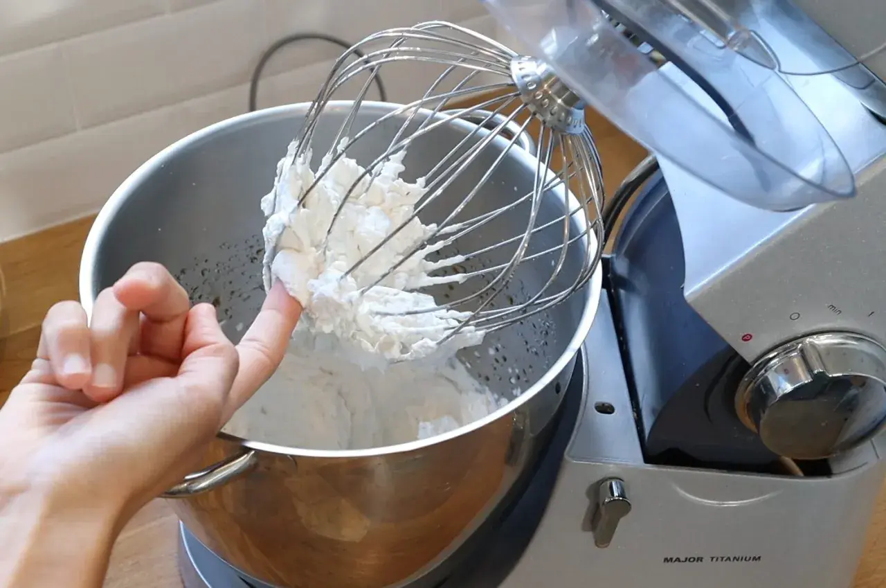 Un doigt teste la texture de la chantilly tranchée sur le fouet d'un robot pâtissier.
